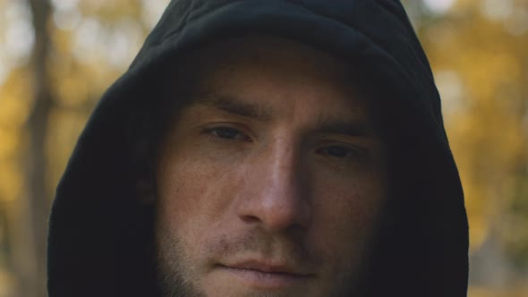 Close Up Portrait of Young Serious Bearded Guy Wearing Hood Looking at Camera at Autumn Park alt