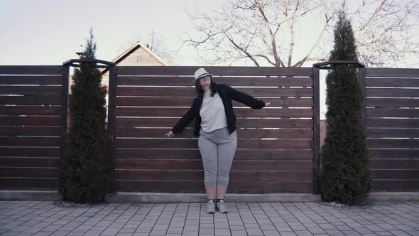 Overweight Woman in a Hat Dancing on the Street Near the Fence in the Village alt