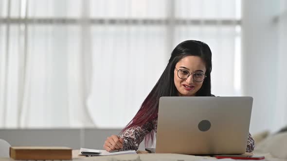 Asian woman sits in office and communicates with client on  video call on laptop alt