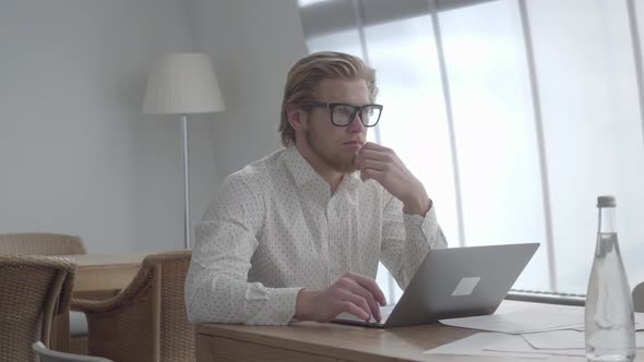 Blond Thoughtful Man in Glasses Sitting at the Table in a Light Comfortable Office in Front alt