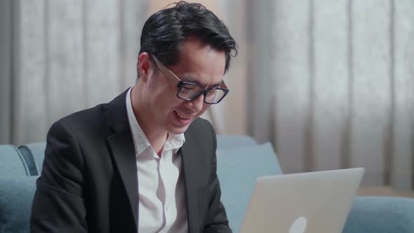 Close Up Of Smiling Asian Businessman In Jacket And Shorts Sitting On Sofa And Typing On A Laptop alt