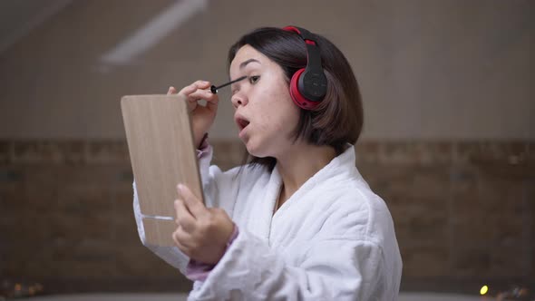 Concentrated Young Little Woman Applying Mascara Looking at Hand Mirror Standing in Bathroom alt
