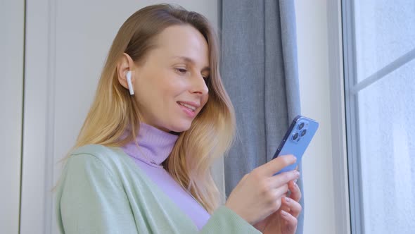 Cheerful blonde woman using mobile phone and listening to favorite music at home in 4k alt