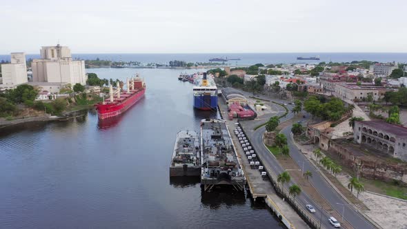 Flight above Ozama River by seaside road traffic towards large ships in downtown harbor port by blue alt