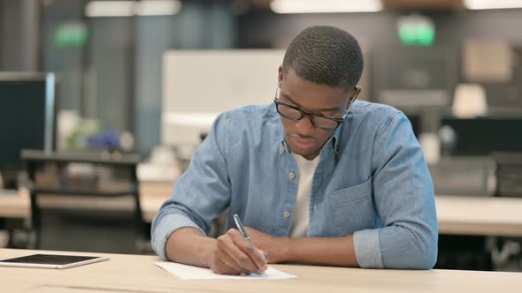 Hardworking Young African American Man Writing on Paper in Office alt