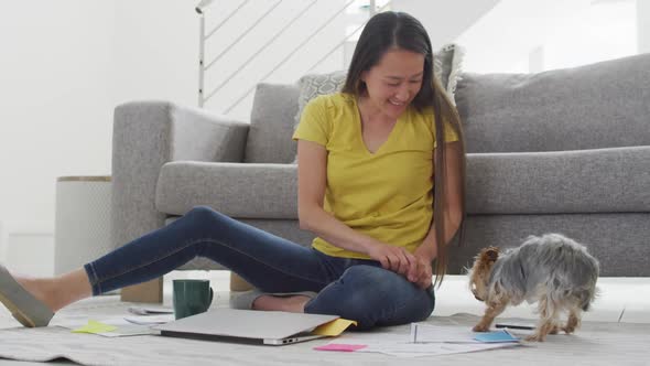 Happy asian woman sitting on floor, working remotely from home with laptop, petting dog alt