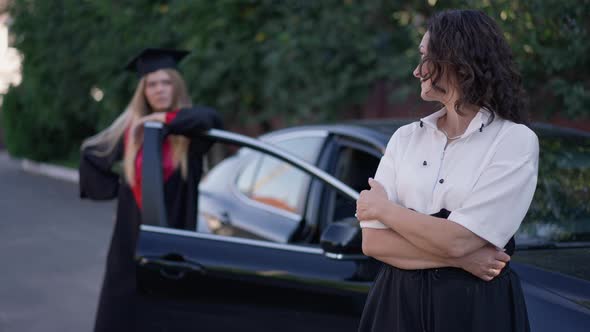 Smiling Mature Mother Looking Back at Blurred Graduate Daughter in Gown Standing at Car alt