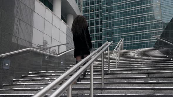 A Brunette with African Curls in a Furry Black Fur Coat Climbs the Stairs Against the Backdrop of alt