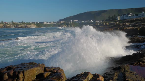 Panning shot of waves crashing violently into rocky coastline of Hermanus, South Africa. alt