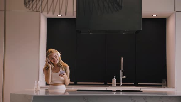 A Young Woman Dancing with a Phone in Her Hands and Headphones in the Kitchen alt