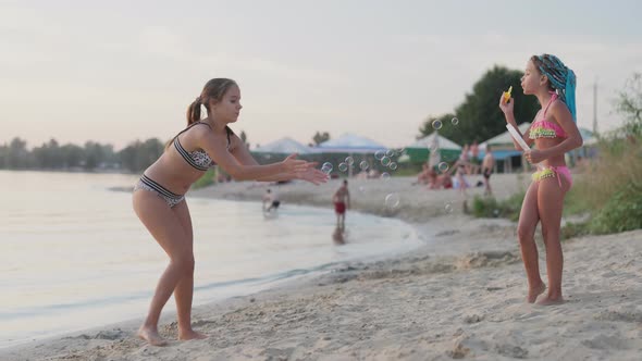 Young Girl Catches Bubbles on the Shore of the Lake Which Her Little Sister Lets Go alt