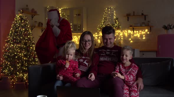 Santa Claus Putting Gifts Under Christmas Tree in Room with Family