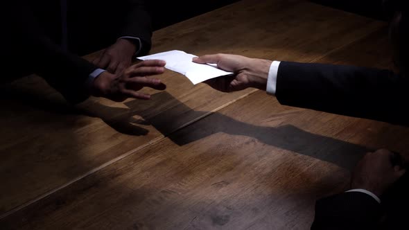 Businessman giving bribe money in white envelope to partner in dark room