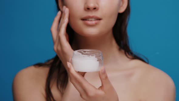 Girl with Wet Hair Holding Moisurizer and Applying It on Her Face Isolated on Blue Background alt