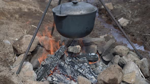 Large Black Cast-Iron Pot Hangs on Tripod Above Burning Bonfire on Field Kitchen