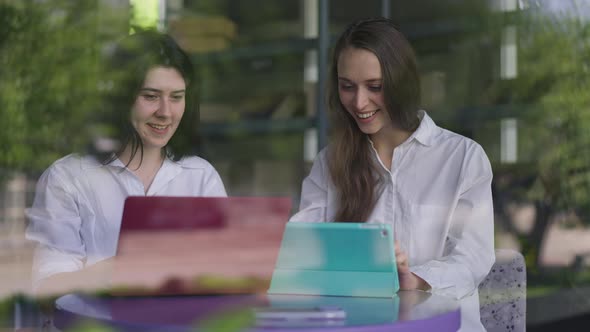 Excited Women Gesturing Victory and Highfive Sitting in Cafe Surfing Internet on Tablet and Laptop alt