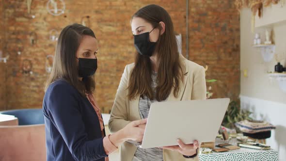 Caucasian female business owner and her coworker wearing face masks, using laptop and talking alt