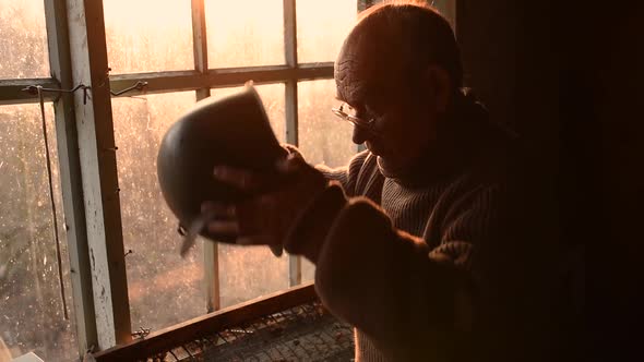 Elderly Man with Glasses Picks Up Military Helmet in Dust and Putting Helmet on Head in House Window alt