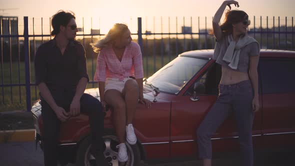 Friends Sit on the Hood of a Car Talk and Laugh at Sunset alt