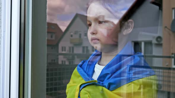 Little Girl with the Flag of Ukraine on Her Shoulders Looks Out the Window with a Serious Expression