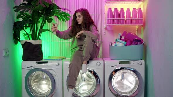 Young Attractive Woman Blogger with Long Curly Hair in Sporty Beige Jumpsuit Sit on Washing Machine alt