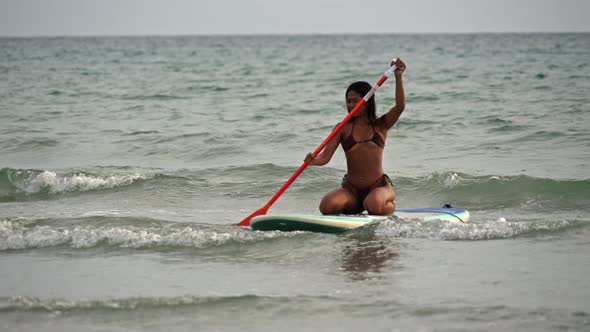 Young Woman In Bikini Kneeling On Paddleboard And Rowing In Sea alt