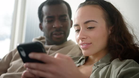 Young Smiling Couple Relaxing At Home On Sofa Checking Social Media On Mobile Phones Together alt