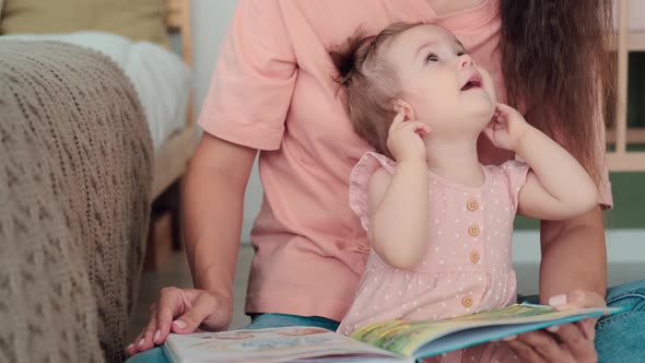 Mom Reads a Book to a Cheerful Kid Sitting on the Floor in the Bedroom alt