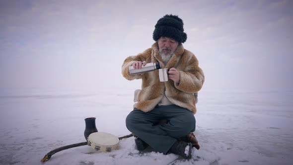Wide Shot Portrait of Old Bearded Mongol Pouring Tea in Cup in Slow Motion Sitting Outdoors on Snowy alt