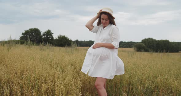 Young Pregnant Woman in White Dress Stands in a Wheat Field Touching Her Belly alt