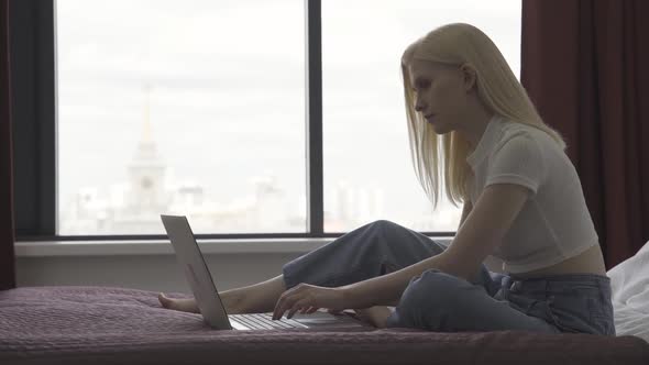 Young Woman Freelancer Works with a Laptop on the Bed Against the Background of a Large Window