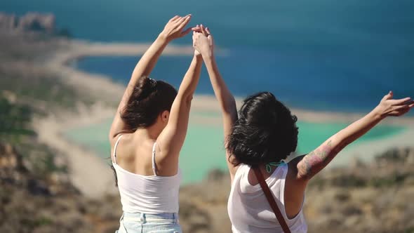 Silhouette of Two Young Girls Standing on Cliff with the View on Blue Sea Lagoon Holding Their Hands alt