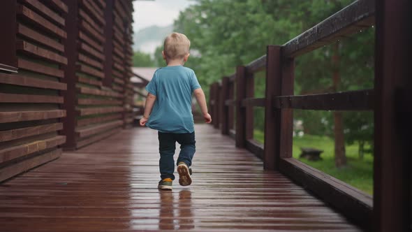 Little Blond Boy Runs Turning Back Along Wet Veranda Deck alt