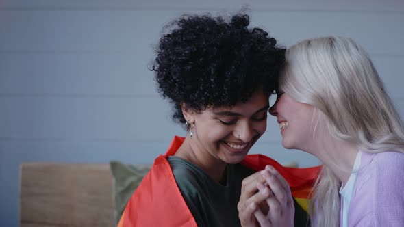 Young Lesbian Couple Kisses Holding Hands Sitting on a Couch Wearing Lgbt Flag alt