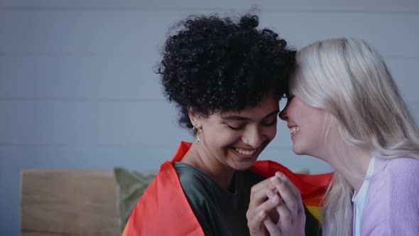 Young Lesbian Couple Kisses Holding Hands Sitting on a Couch Wearing Lgbt Flag