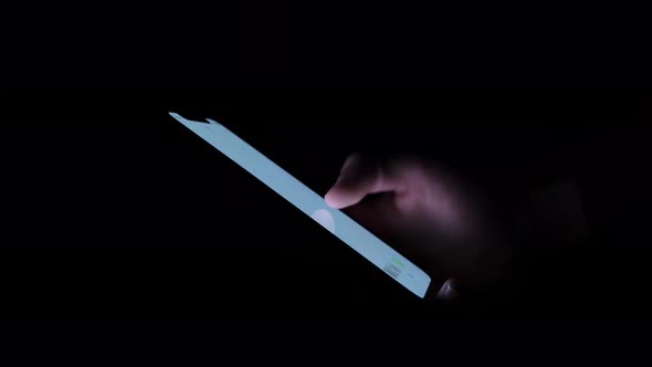 Close-up holding and using a smartphone in a dark room, white glowing screen. alt
