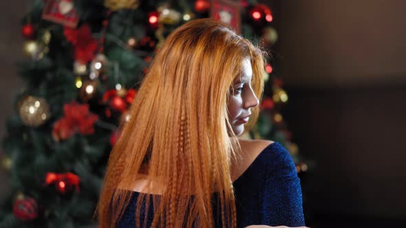 Beautiful Red-haired Woman Looks at Camera Against the Background Christmas Tree alt