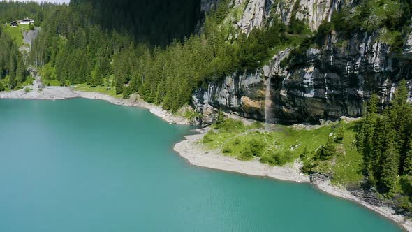 Aerial of hikers walking underneath cliffs with a small waterfall running into a beautiful lake alt