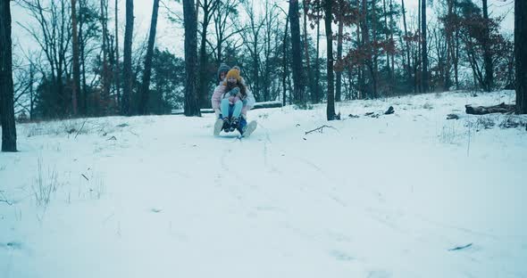 Girl Sits on Mother Laps Sliding Down Snowy Hill on Sled alt