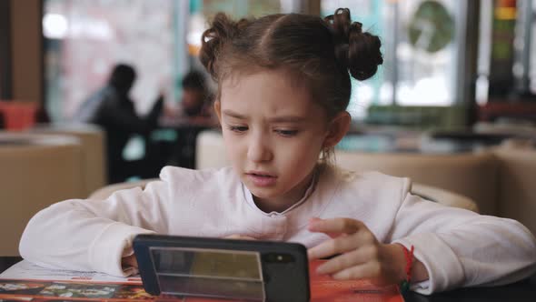 A Little Girl at a Table in a Cafe Sitting with a Smartphone in Hand