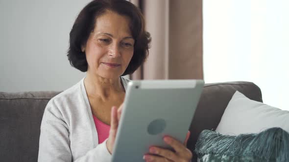 Mature Caucasian Woman Sitting on Sofa with Electronic Tablet in Hand at Home alt