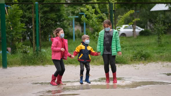 Two little girls and a boy in medical masks and rubber boots jump through puddles