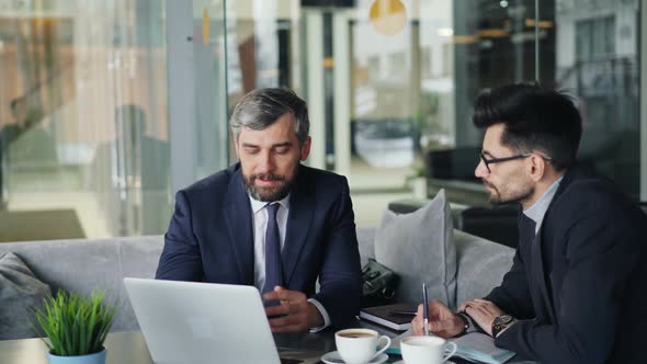 Mature Businessmen Talking in Cafe Looking at Laptop Screen Discussing Contract alt