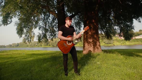 A young guy in a black T-shirt plays an acoustic guitar on a background of a tree and a river.