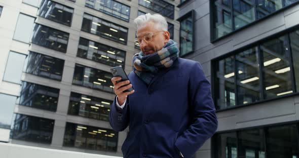 A Grayhaired Man Uses a Mobile Phone Standing in Business Center of the City alt