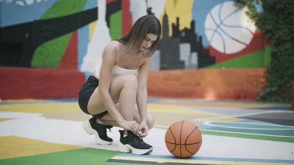 Young Slim Basketball Player Tying Sneakers Shoelaces and Leaving with Orange Ball alt