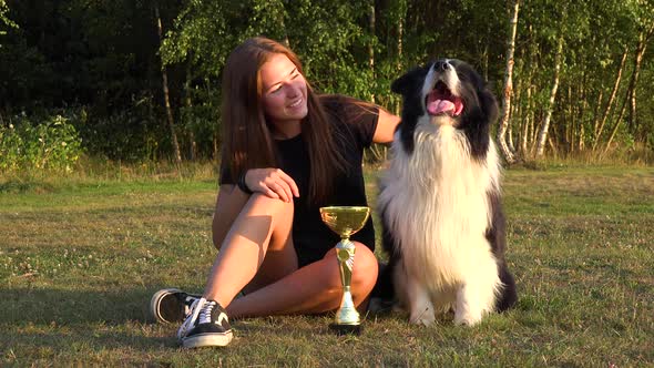 A Woman and a Border Collie Sit in a Meadow with a Trophy, the Woman Shows the Trophy To the Dog alt