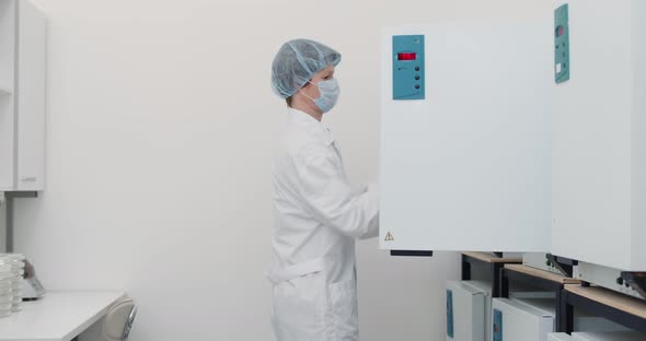 Female Scientist in White Coat Mask and Cap Using Large Fridge Unit in Lab alt