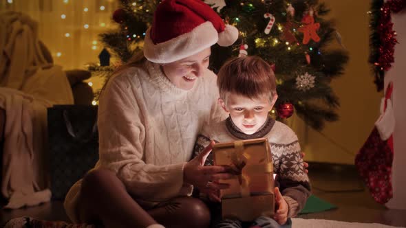Happy Mother Hugging Her Son Unwrapping Christmas Gifts and Present in Living Room alt