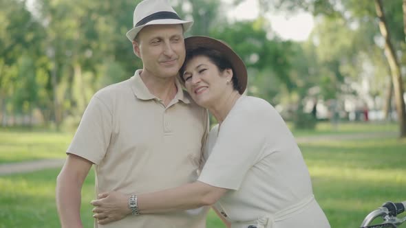 Happy Loving Woman Hugging Husband in Park and Smiling at Camera. Man Looking at Wife with Love alt